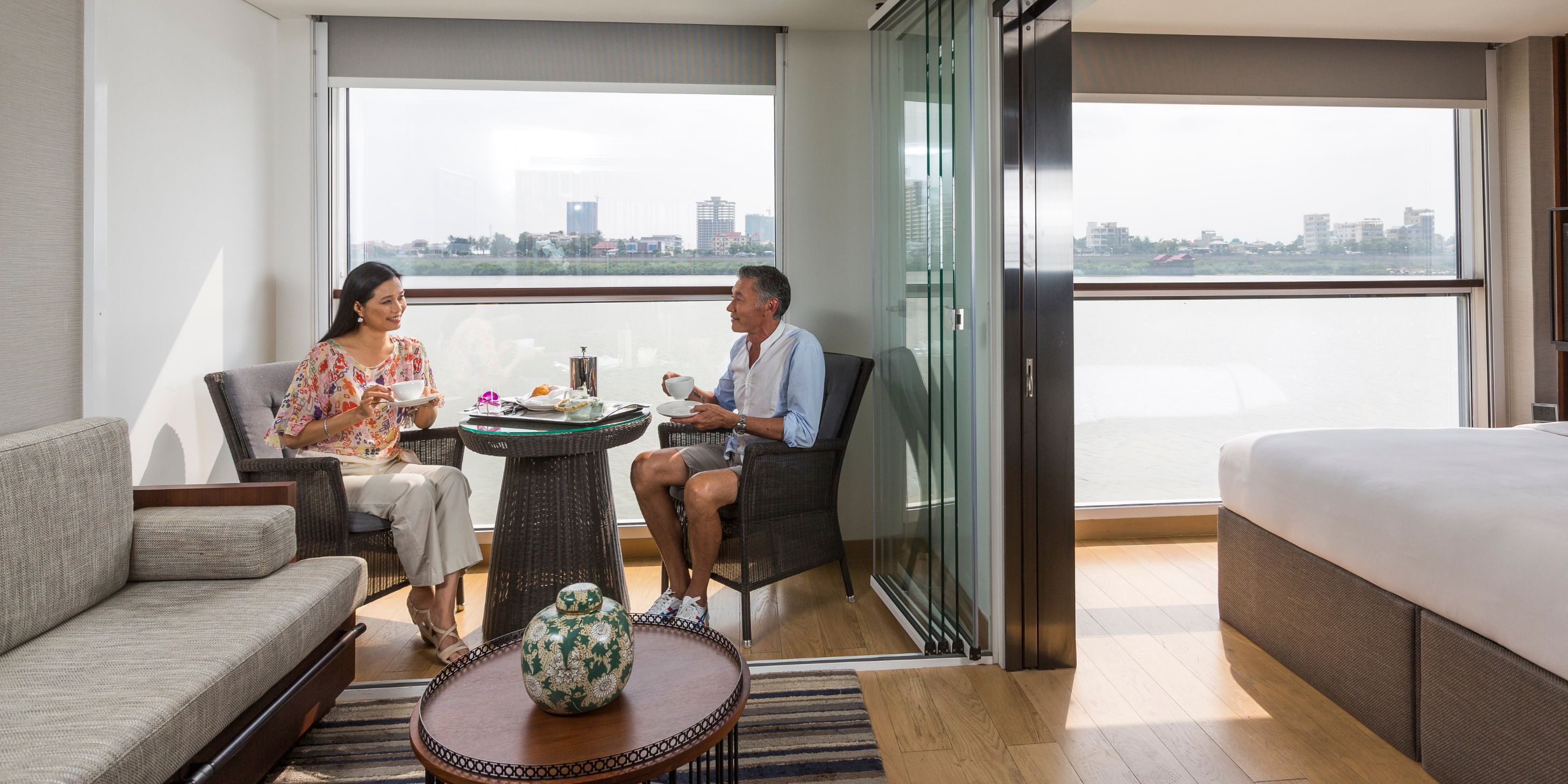two guests sitting at a small dining table in their suite onboard a boutique river cruise with a view of the Mekong river in the background through the windows