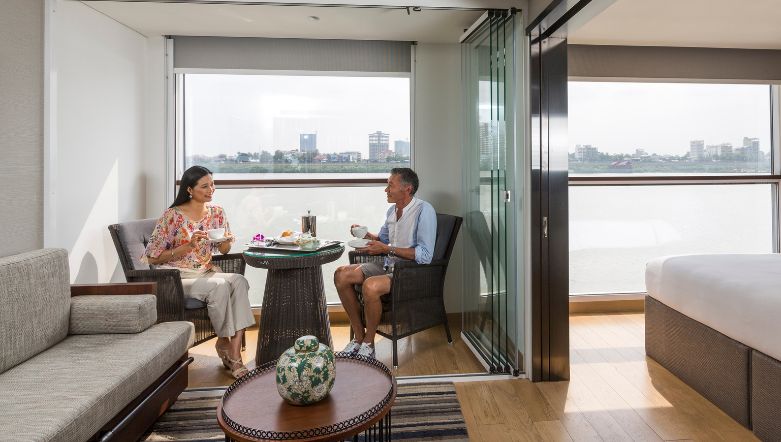 two guests sitting at a small dining table in their suite onboard a boutique river cruise with a view of the Mekong river in the background through the windows